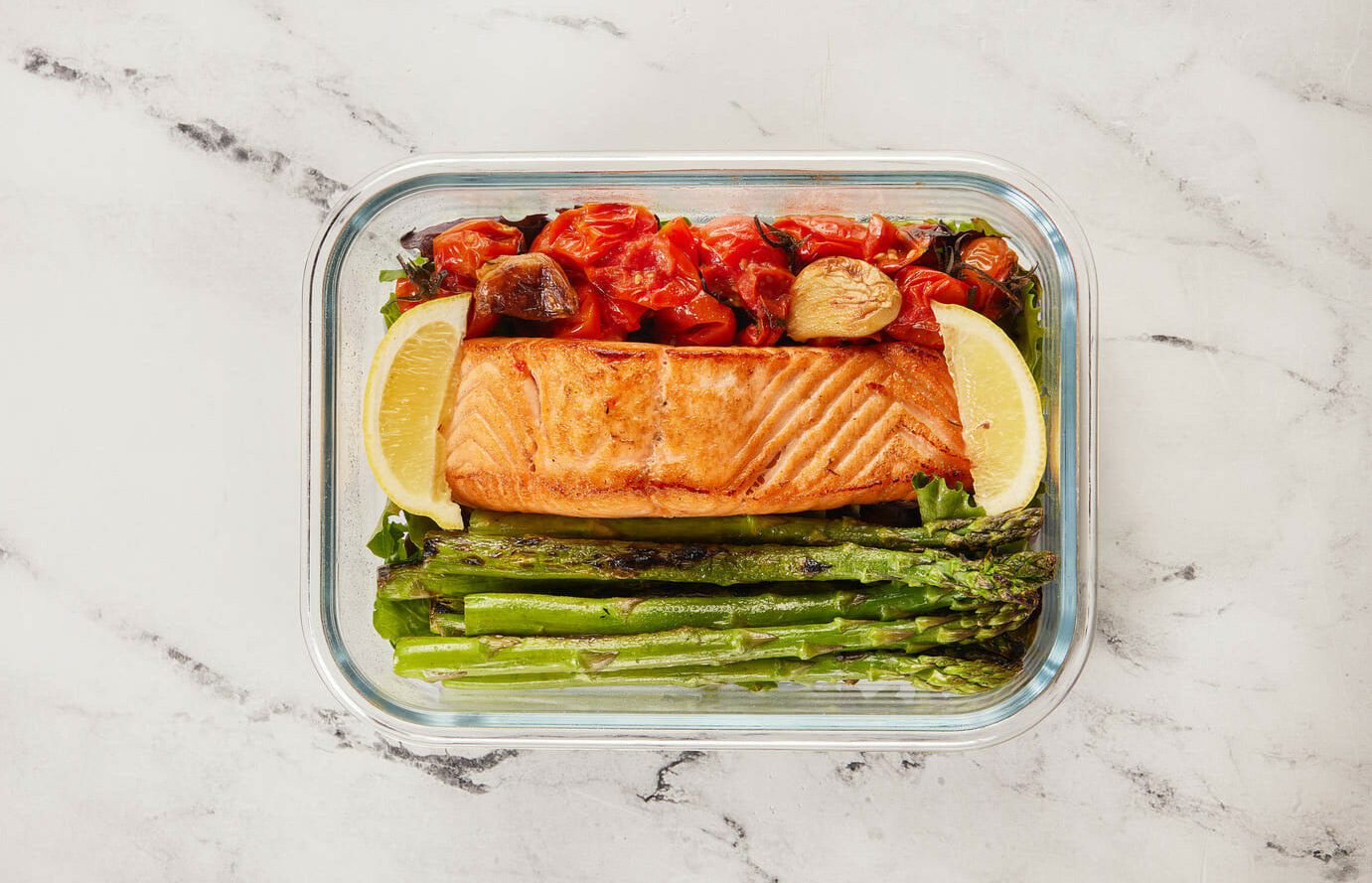 A glass meal-prep container holds a salmon fillet, grilled asparagus, roasted cherry tomatoes, and lemon wedges.